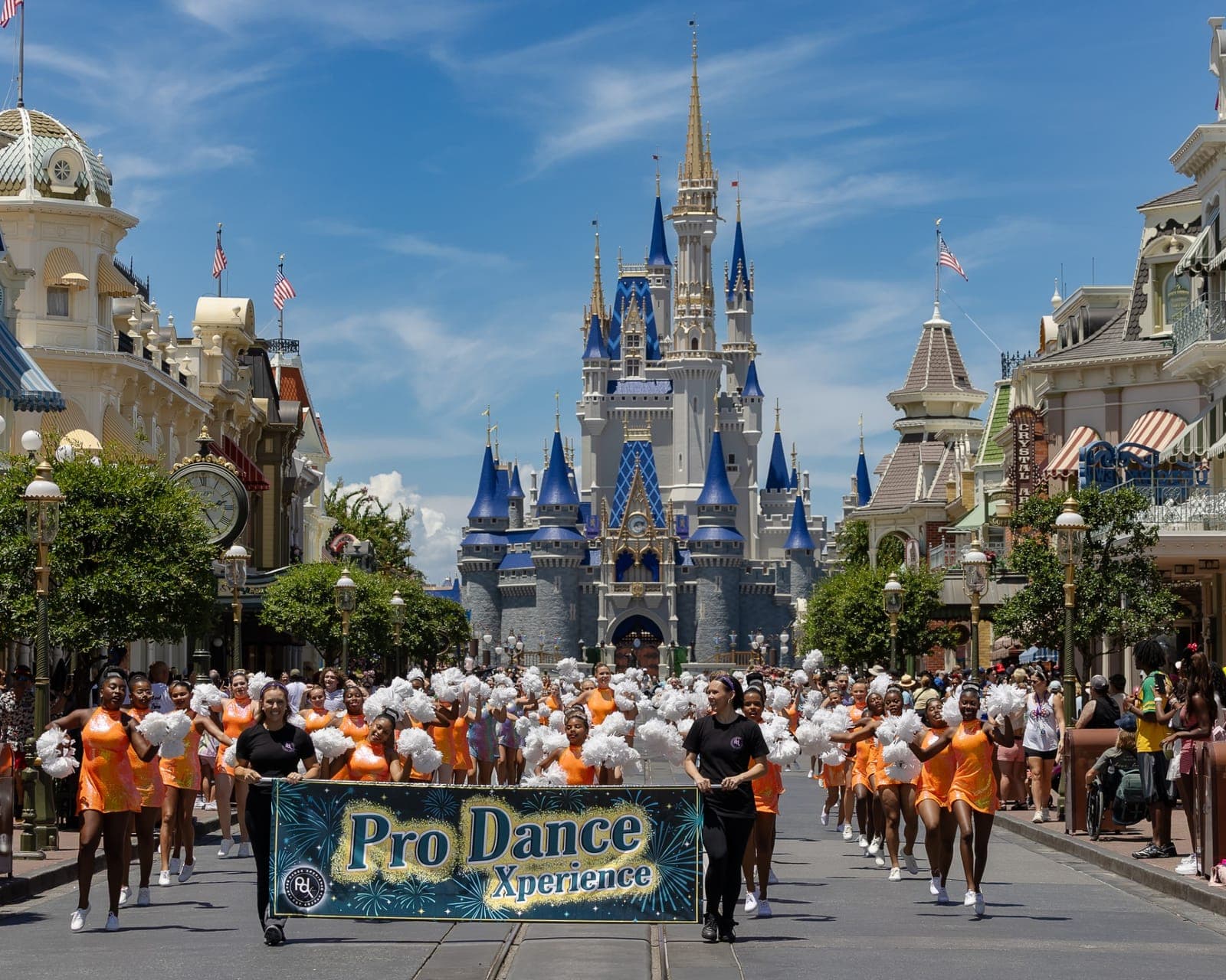 Dancers in orange costumes with white pom-poms march behind a Pro Dance Xperience banner on Main Street, with Cinderella Castle behind them