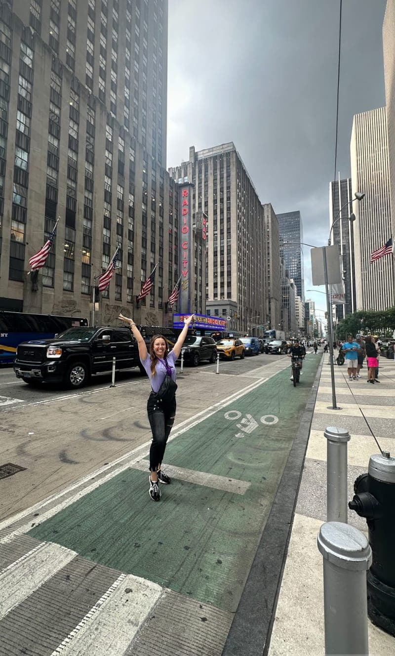 A woman in a purple top raises both arms in celebration in a Midtown Manhattan street, with Radio City Music Hall's sign behind her